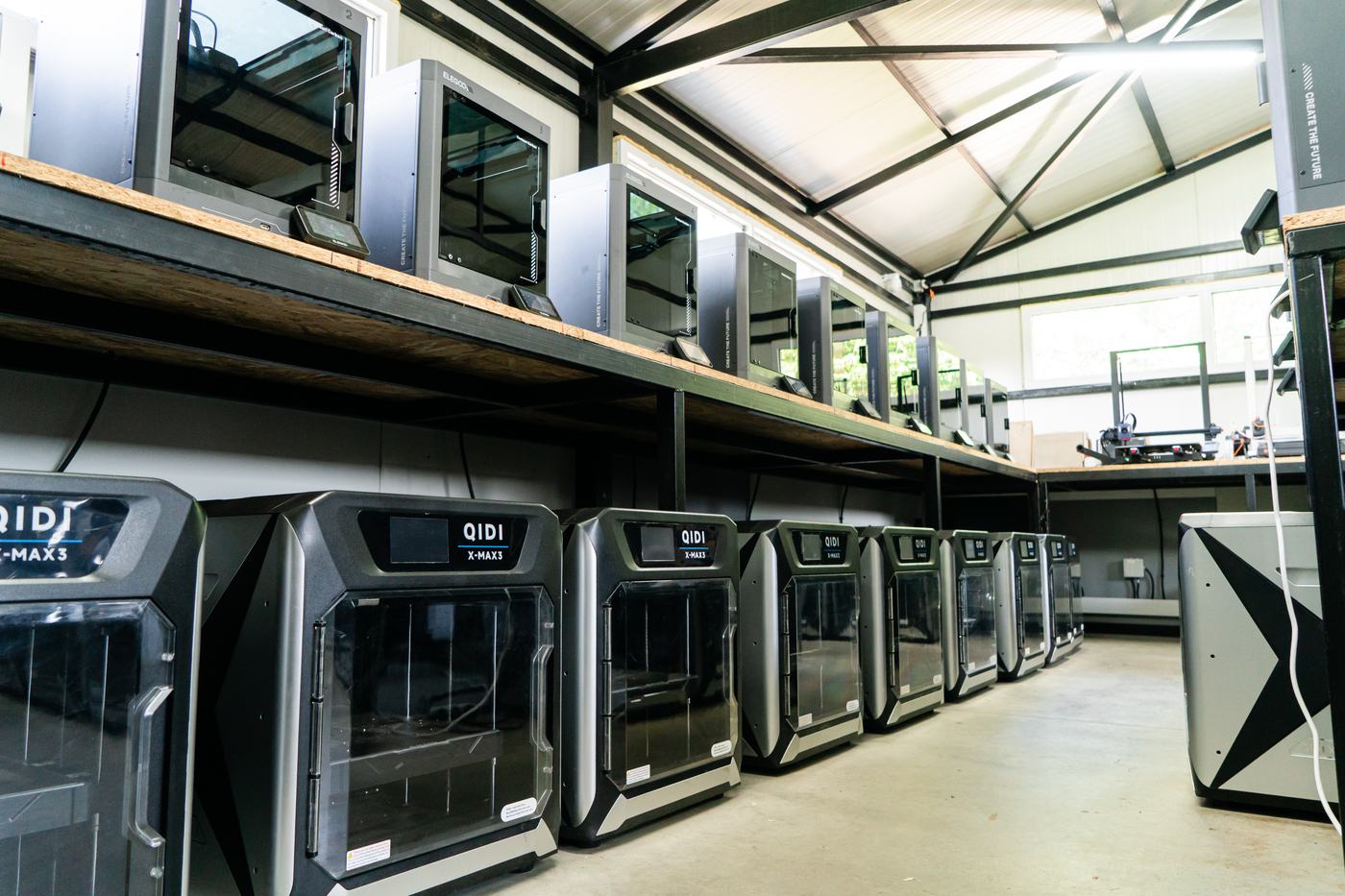 Row of enclosed FDM printers standing at floor level with a further shelf of machines above them, seen down the length of the bench