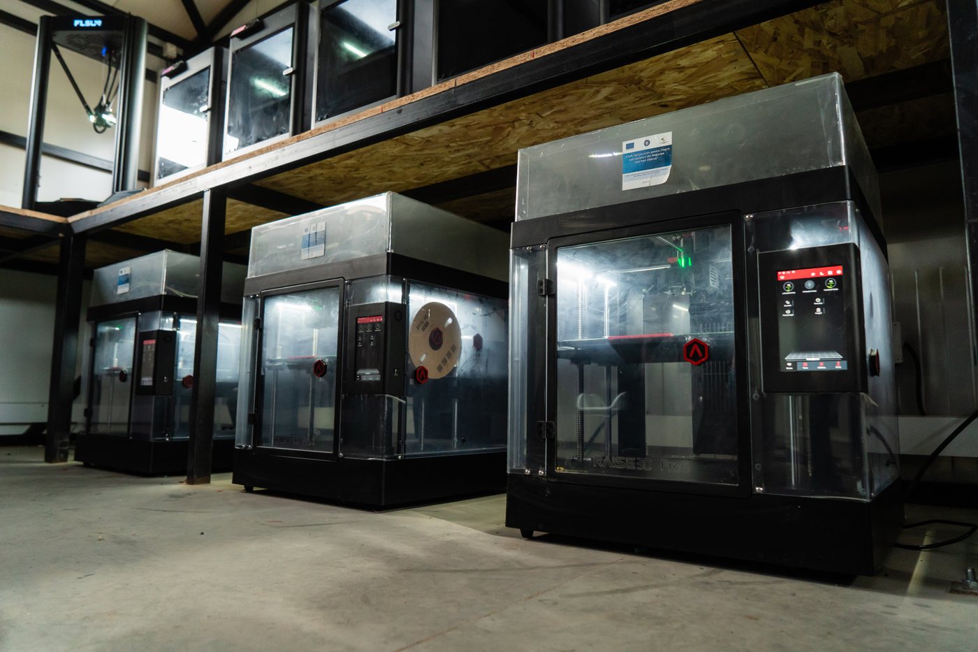 Three industrial FDM printers with clear front panels, lit build chambers and red emergency stop buttons, standing beneath a shelf of further machines