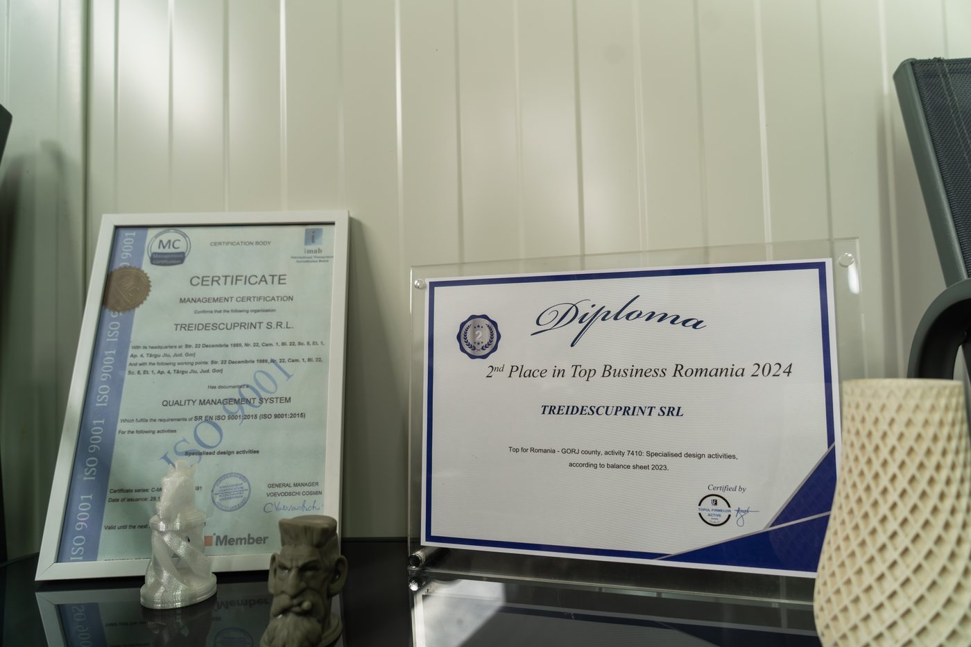Framed ISO 9001 quality management certificate issued to TREIDESCUPRINT S.R.L. beside a 2nd Place in Top Business Romania 2024 diploma, with three printed sample parts in front of them