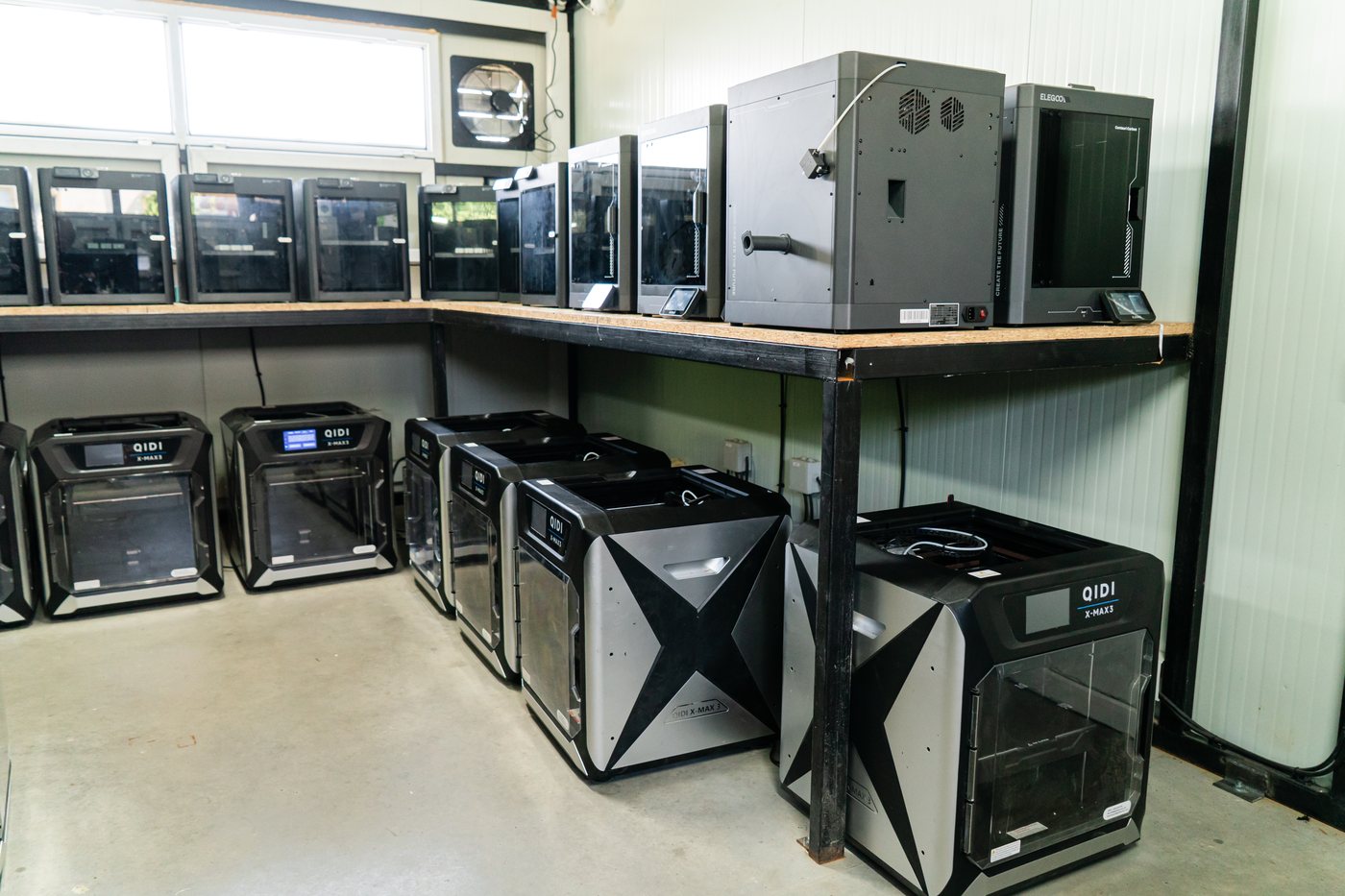 Corner of the production hall with enclosed FDM printers on two bench decks and a wall-mounted extraction fan above them
