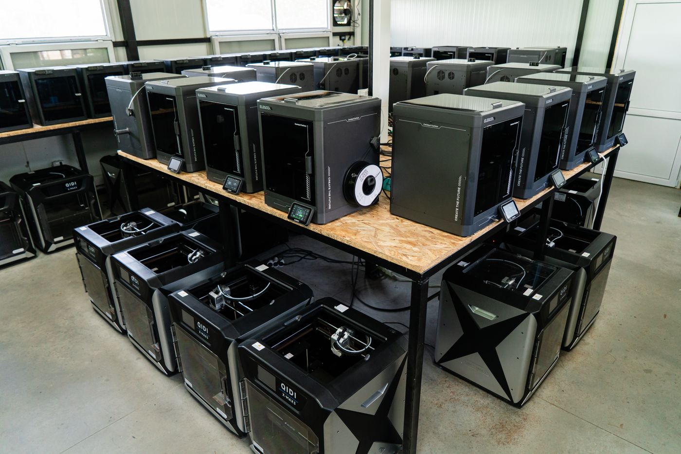 Two decks of enclosed FDM printers filling an L-shaped bench island, with more machines along the wall behind