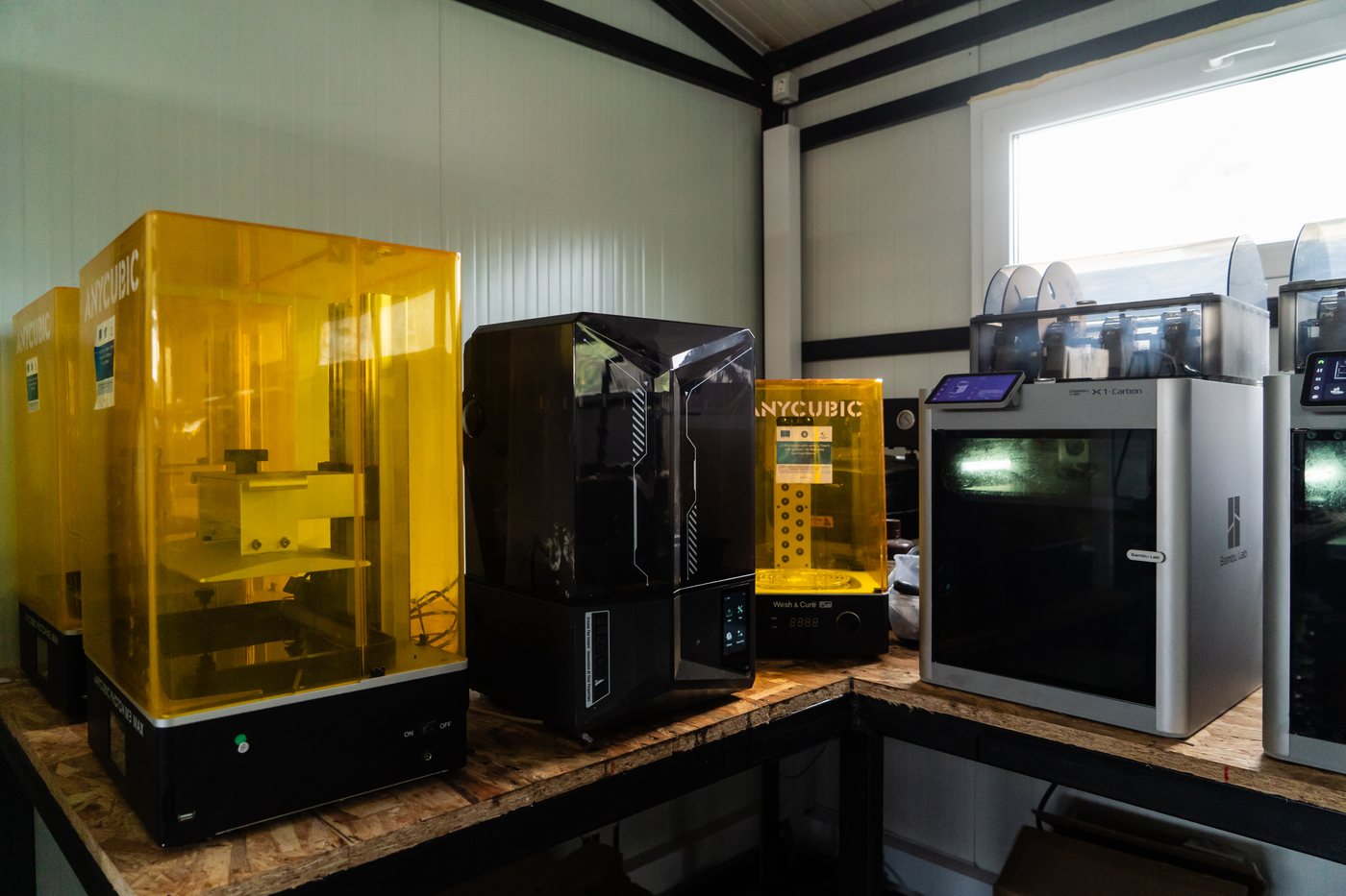 Two large-format resin printers with yellow UV filtering covers beside a wash and cure station on a workshop bench